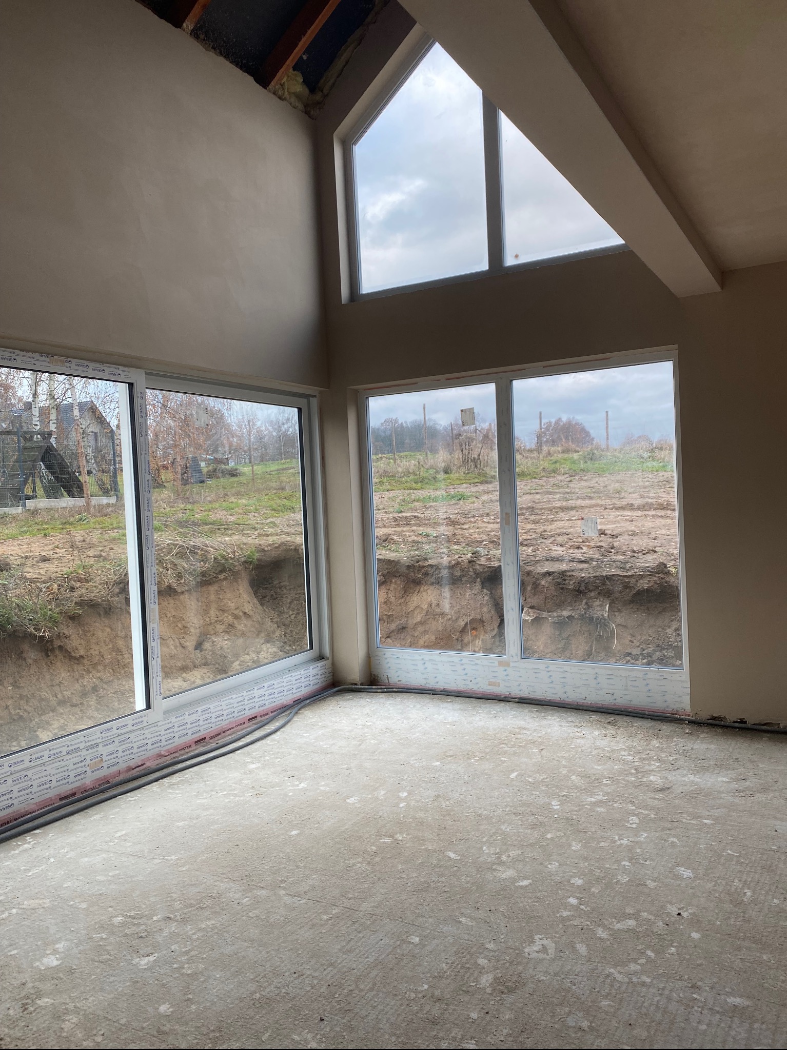 Wnętrze surowego domu po nałożeniu gładzi gipsowej na ścianach, widoczne duże okna z widokiem na wykopany teren.