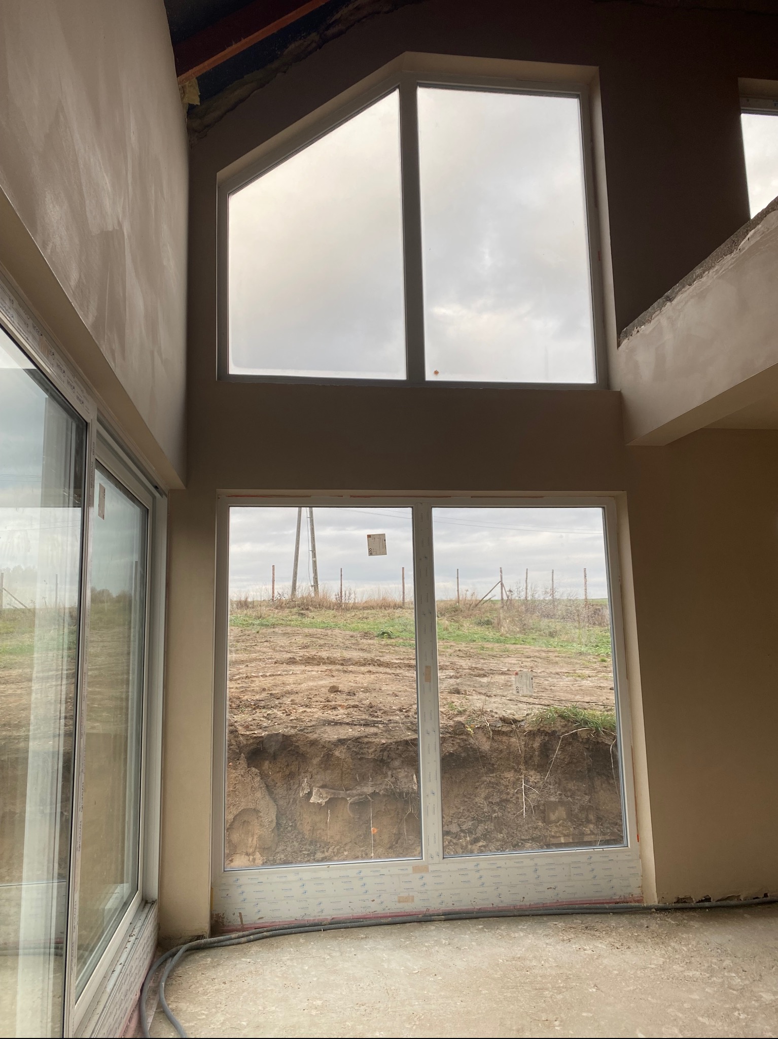 Wnętrze w stanie surowym zamkniętym z oknami i świeżo położonymi tynkami gipsowymi na ścianach, widok na wykop ziemny za oknem.