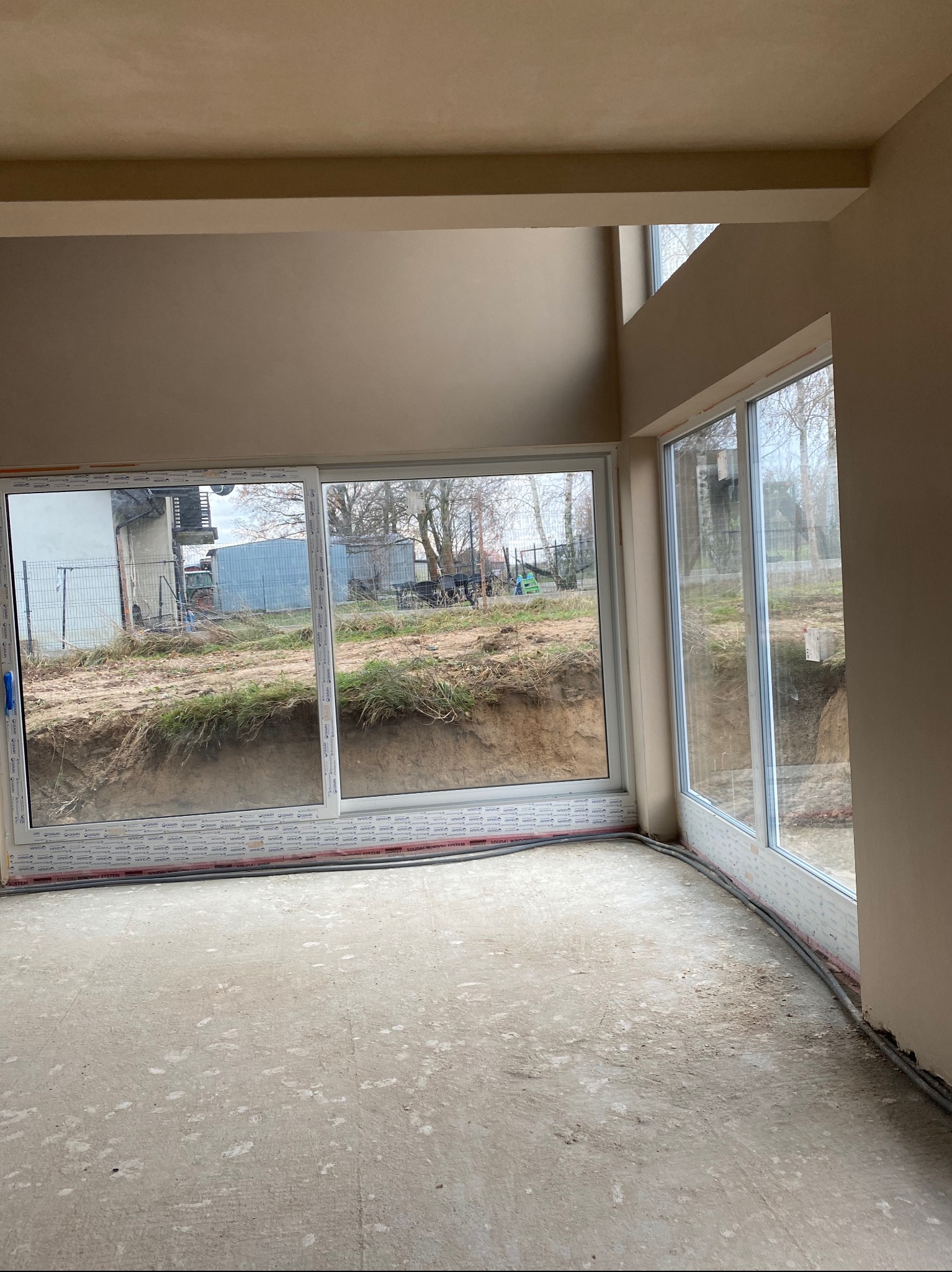 Wnętrze w stanie deweloperskim po nałożeniu gładzi szpachlowej na ścianach, z widocznymi dużymi oknami przesuwnymi i przewodami elektrycznymi przy podłodze.