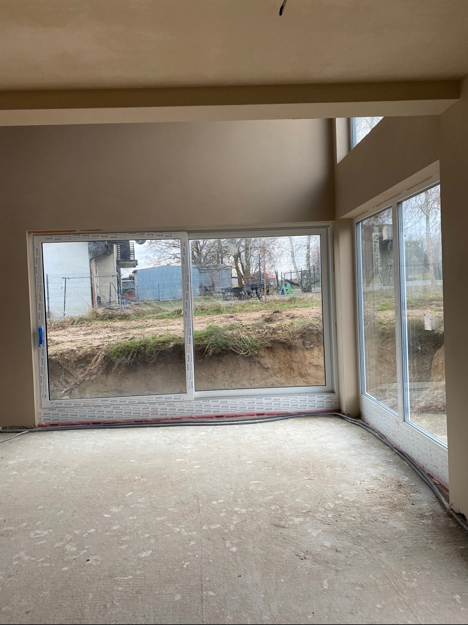 Wnętrze surowego domu po tynkowaniu maszynowym, z widocznymi oknami i przygotowaną podłogą, ściany w kolorze beżowym, widok na ogród przez okna.