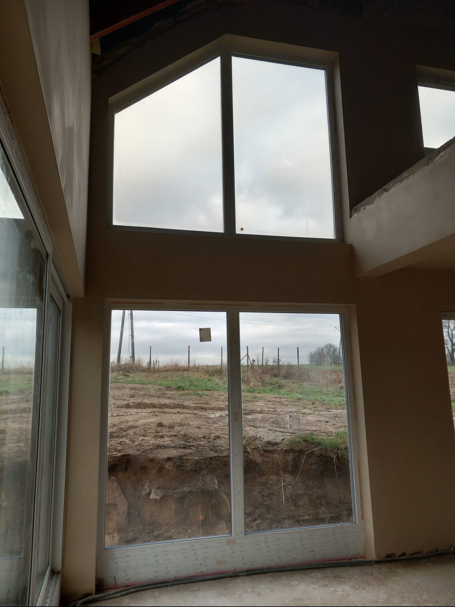 Wnętrze domu w trakcie prac wykończeniowych, widoczne ściany po nałożeniu gładzi gipsowej i duże okna z widokiem na krajobraz za oknem.