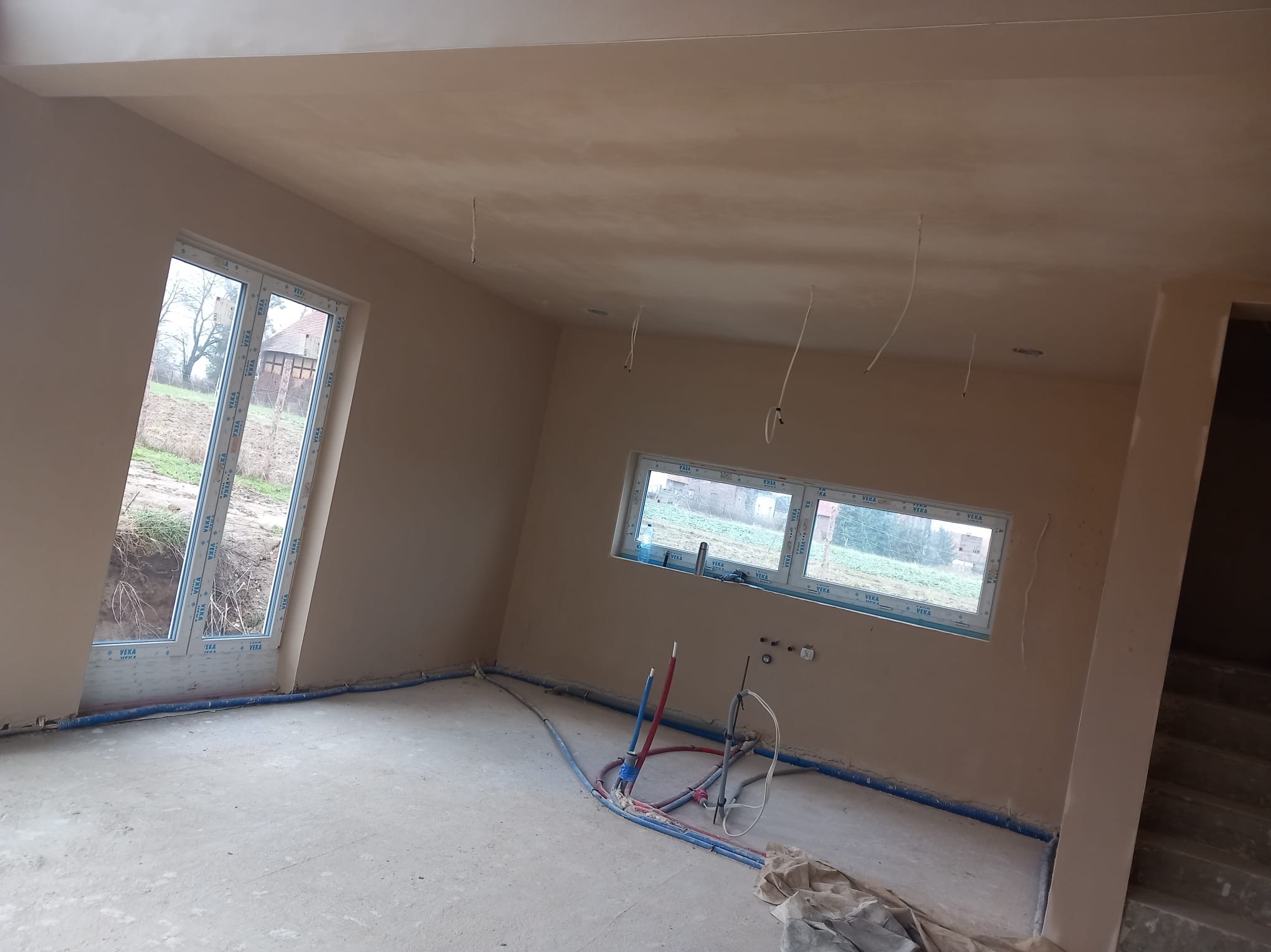 Wnętrze w trakcie prac wykończeniowych: świeżo otynkowane ściany w odcieniach beżu, widoczne instalacje hydrauliczne i elektryczne, okna PCV z widokiem na zewnątrz, surowy beton na podłodze.