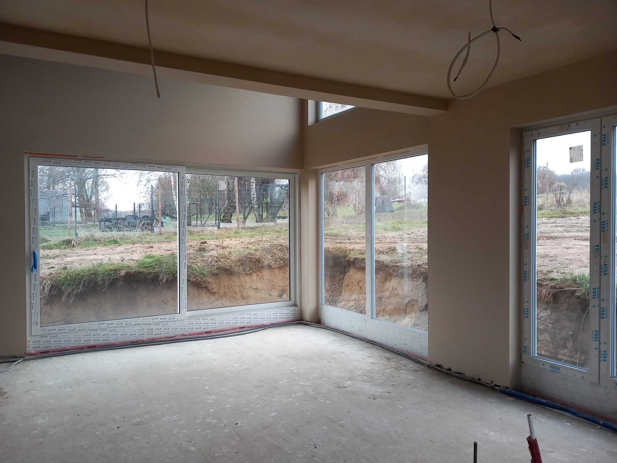 Surowe wnętrze z oknami PCV i świeżo nałożonymi, jasnymi tynkami na ścianach, widoczne niedociągnięcia na podłodze i wystające kable z sufitu, przez okna widok na wykop ziemny.