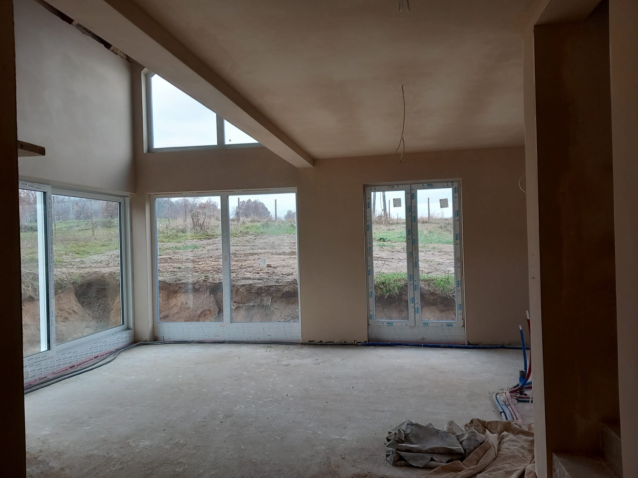 Wnętrze domu w trakcie prac wykończeniowych, ściany pokryte świeżym jasnym tynkiem, duże okna z widokiem na wykop ziemny, widoczne instalacje na podłodze.