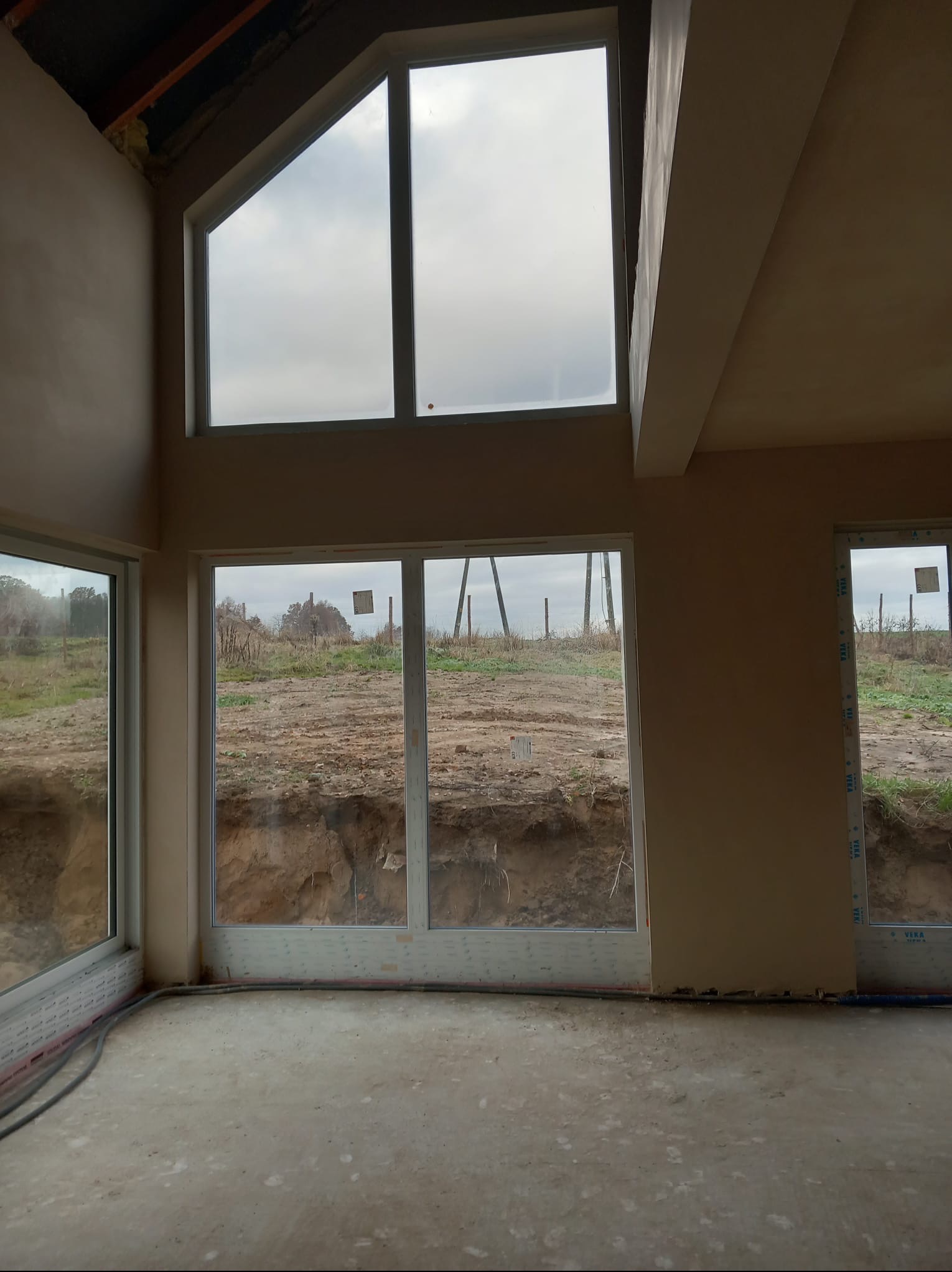 Wnętrze domu w stanie surowym po nałożeniu tynków, widoczne okna z widokiem na wykop ziemny, surowe ściany i podłoga.