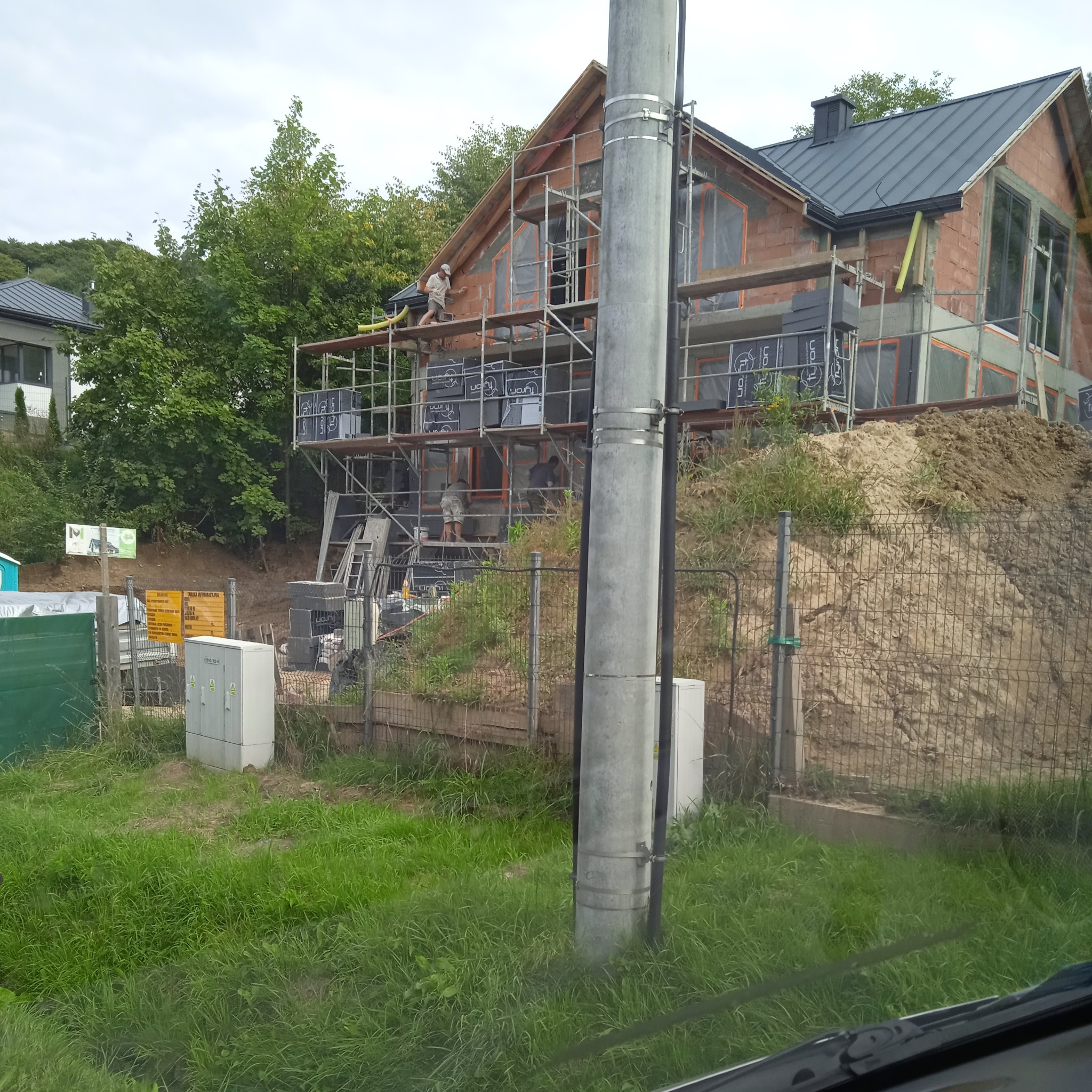 Dom w trakcie ocieplania, widoczne rusztowania, warstwa izolacyjna z nadrukiem, mur z czerwonej cegły oraz pracownicy na rusztowaniu.