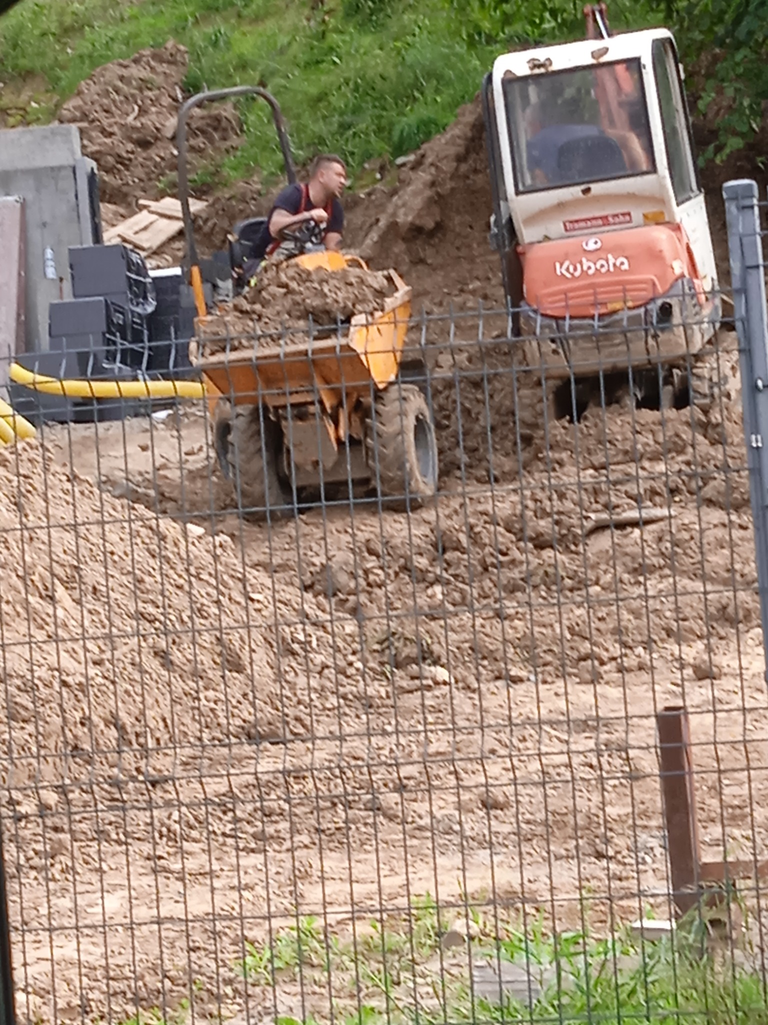 Operator w małej wywrotce z ładunkiem ziemi, obok koparka Kubota na placu budowy, widok przez metalowe ogrodzenie.