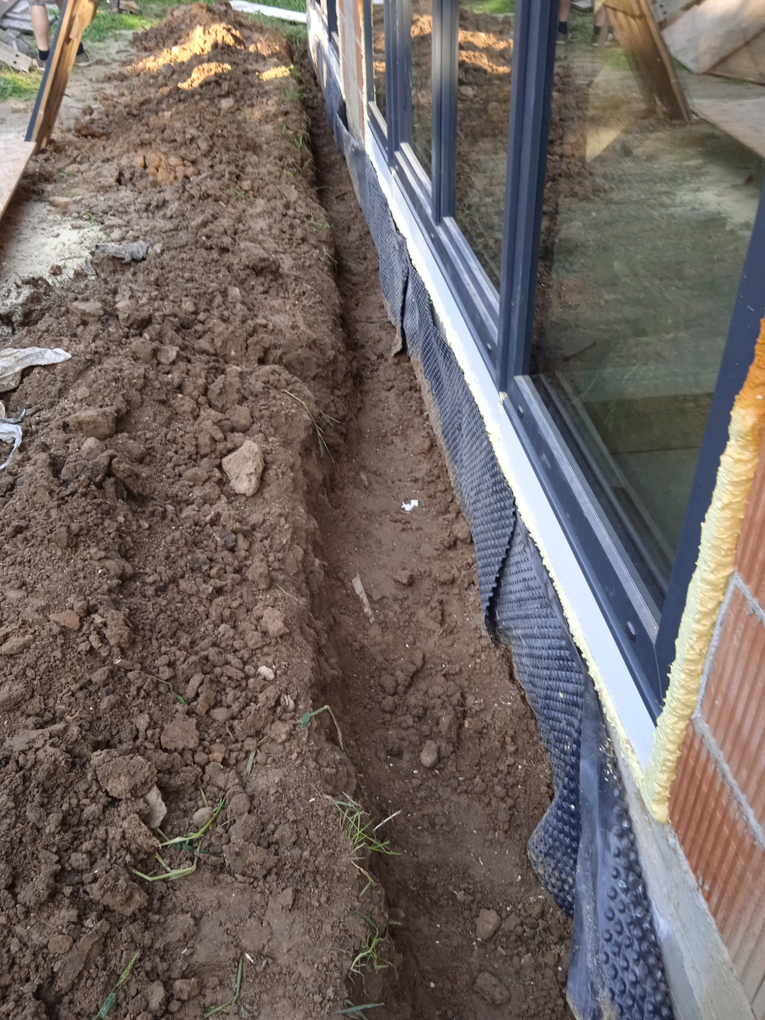 Wykop w ziemi wzdłuż ściany budynku z oknami, zabezpieczonej folią kubełkową i pianką montażową, prawdopodobnie przygotowanie do ocieplenia fundamentów.