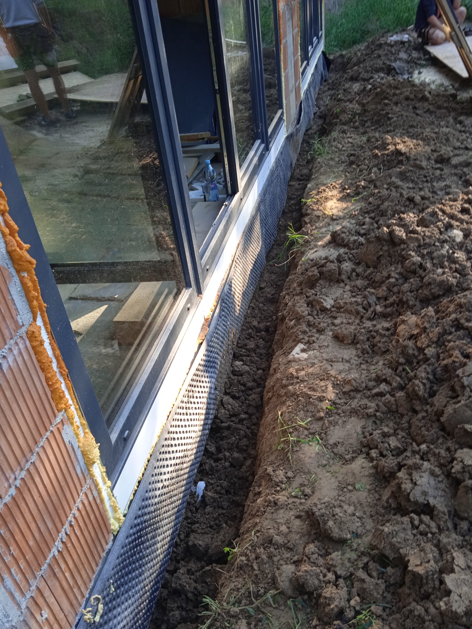 Fundament budynku z izolacją pionową z folii kubełkowej, widoczny wykop ziemny i fragment ściany z cegły z pianką montażową.