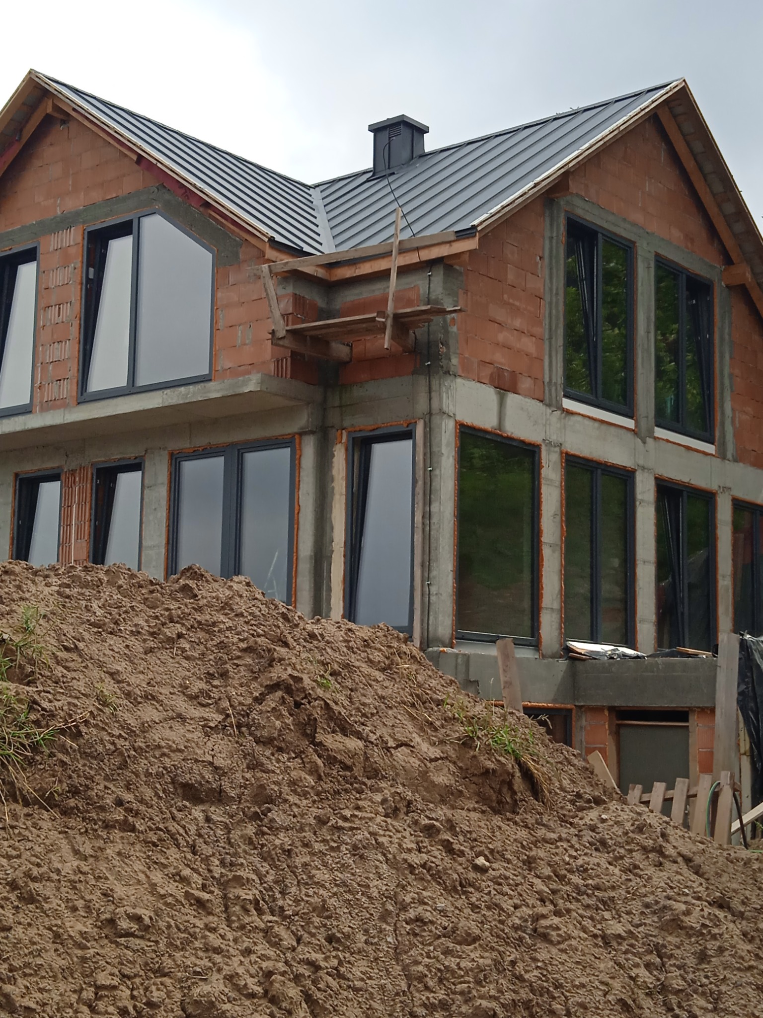 Surowy stan budynku z cegły z zamontowanymi oknami, widoczna sterta ziemi przed budynkiem, element konstrukcji elewacji w trakcie prac.