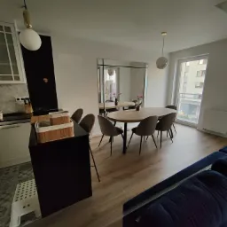 Nowoczesne wnętrze mieszkania z drewnianą podłogą, owalnym stołem z krzesłami, czarnym aneksem kuchennym i oknem z widokiem na budynek mieszkalny.