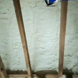 Poddasze z drewnianą konstrukcją pokryte grubą warstwą jasnej, spienionej izolacji, widoczny fragment logo firmy w rogu