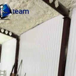 Wnętrze magazynu z metalowymi ścianami i stropem pokrytym białą pianką izolacyjną, widoczne brązowe elementy konstrukcyjne oraz logo firmy wykonującej prace w rogu.