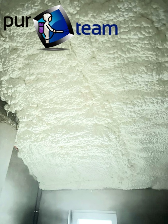 Świeżo nałożona, jasna pianka izolacyjna pokrywa sufit i fragment ściany, widoczny także fragment okna oraz logo firmy w lewym górnym rogu.