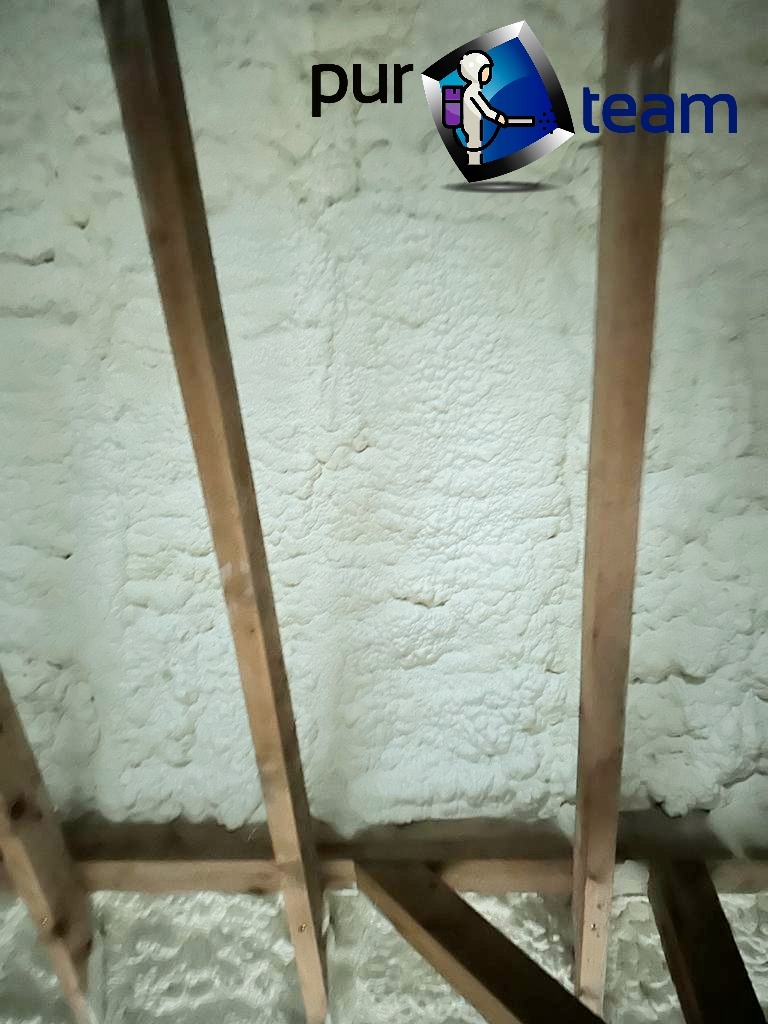 Poddasze z drewnianą konstrukcją pokryte grubą warstwą jasnej, spienionej izolacji, widoczny fragment logo firmy w rogu