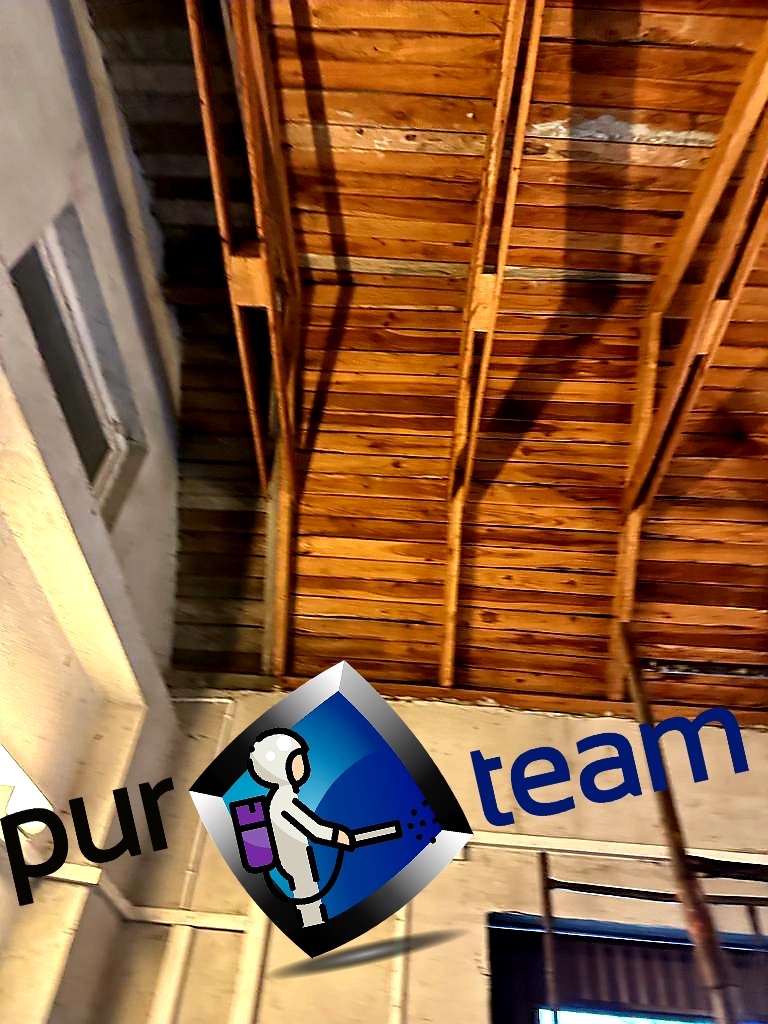 Zdjęcie przedstawia drewnianą konstrukcję dachu od wewnątrz, z widocznymi belkami i deskami, oraz fragment ściany z oknem. W rogu znajduje się logo firmy zajmującej się natryskiem izolacji.