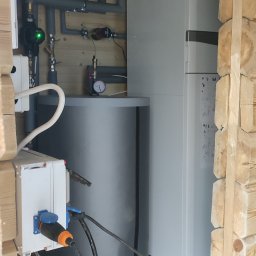 Kornel Rzepkowski Hydro-Therm - Wnętrze kotłowni z zainstalowaną pompą ciepła i zbiornikiem buforowym, widoczne rury instalacji grzewczej, elektryka w trakcie podłączenia, budynek w trakcie wykończenia.