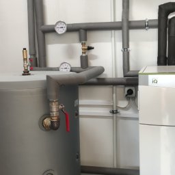 Kornel Rzepkowski Hydro-Therm - Szary zbiornik buforowy z zaworem i manometrem obok białej pompy ciepła Vaillant Green iQ z czarnymi rurami izolowanymi, zamontowane w pomieszczeniu technicznym.