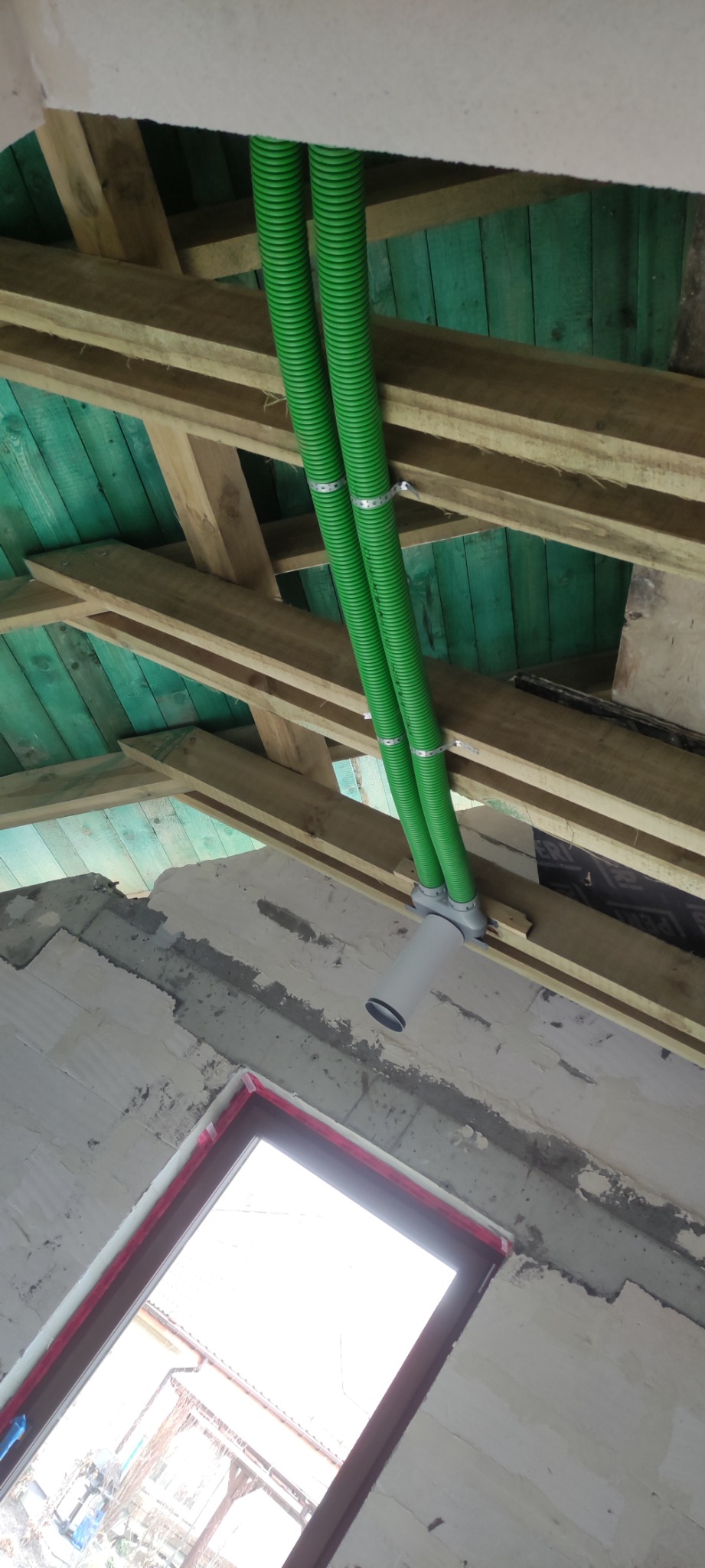 Dwa zielone, karbowane przewody wentylacyjne przymocowane metalowymi obejmami do drewnianej konstrukcji dachu z zieloną podbitką, zakończone białym elementem instalacji wentylacyjnej w surowym...