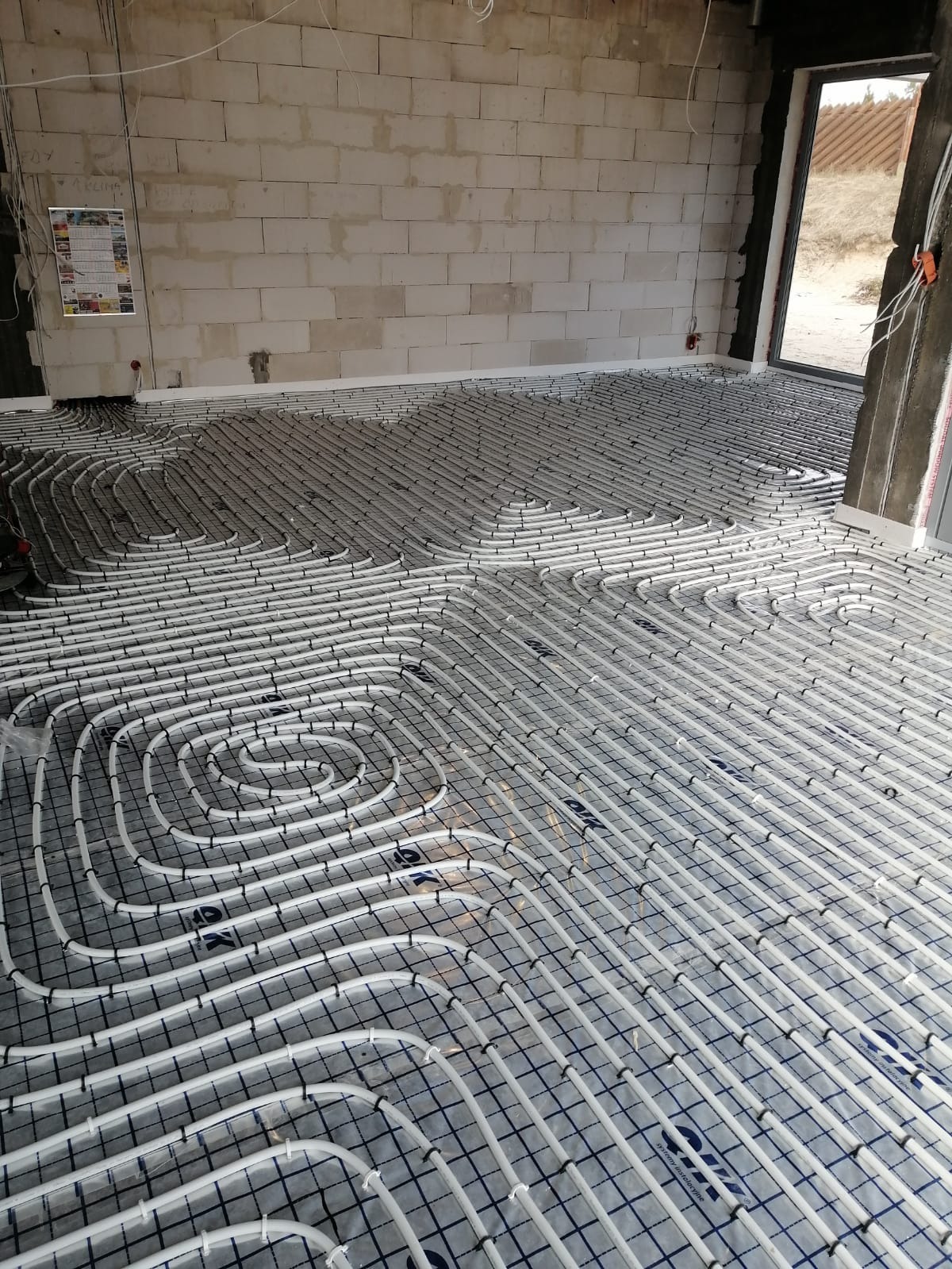 Rozłożone rury ogrzewania podłogowego na folii z siatką w surowym wnętrzu, widoczne ściany z betonu komórkowego i okno.