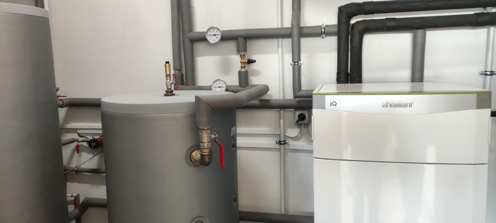 Szary zbiornik buforowy z zaworem i manometrem obok białej pompy ciepła Vaillant Green iQ z czarnymi rurami izolowanymi, zamontowane w pomieszczeniu technicznym.