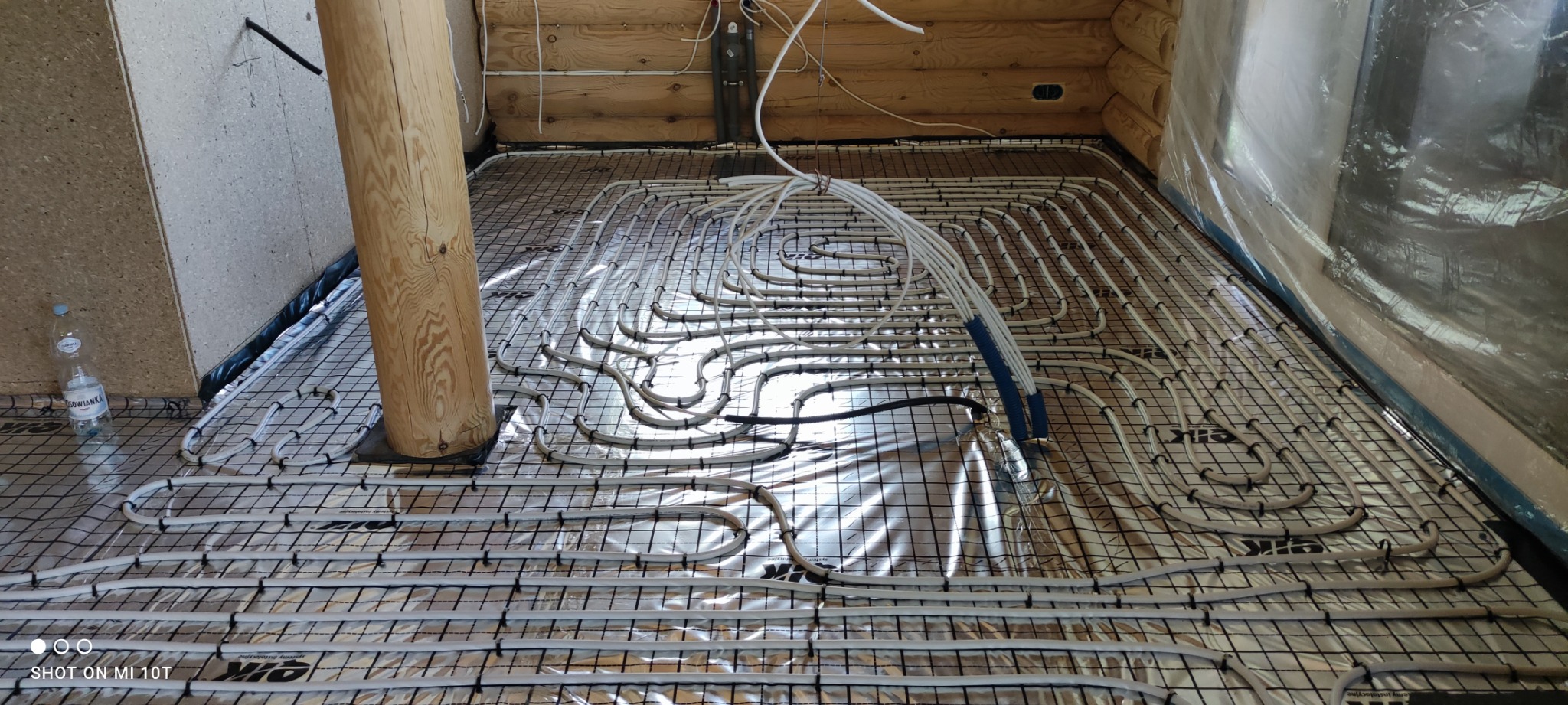 Rozłożona instalacja ogrzewania podłogowego na folii aluminiowej, rury mocowane do siatki zbrojeniowej, widoczny drewniany słup i butelka wody mineralnej.