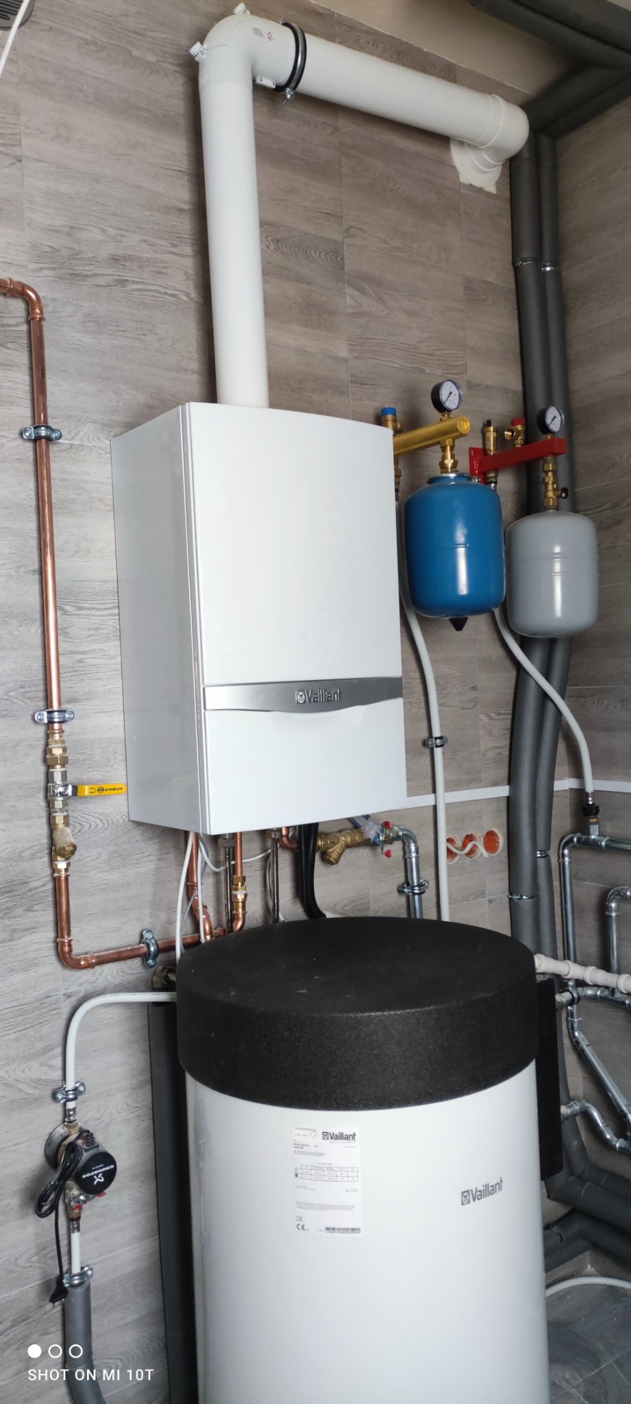 Biały piec gazowy Vaillant z podłączonym zasobnikiem ciepłej wody, rurami miedzianymi i elementami instalacji grzewczej na tle płytek imitujących drewno.