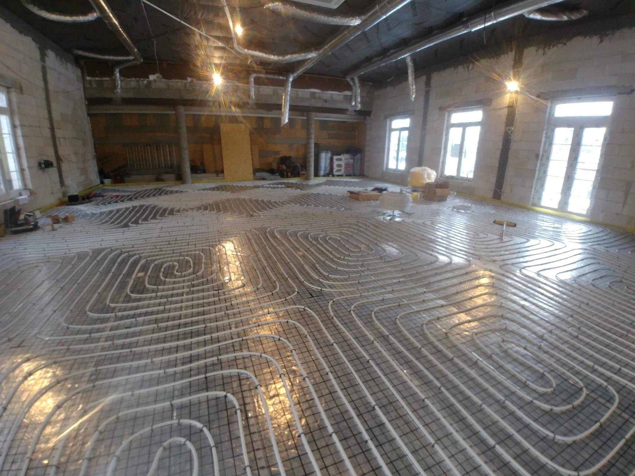 Rozłożone rury ogrzewania podłogowego na folii izolacyjnej z siatką w surowym wnętrzu z oknami, widoczne elementy instalacji pod sufitem.