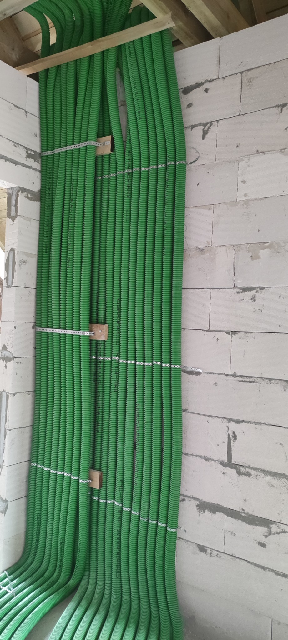 Równolegle ułożone, zielone rury karbowane przymocowane do ściany z białych bloczków betonowych za pomocą metalowych uchwytów i drewnianych elementów dystansowych.