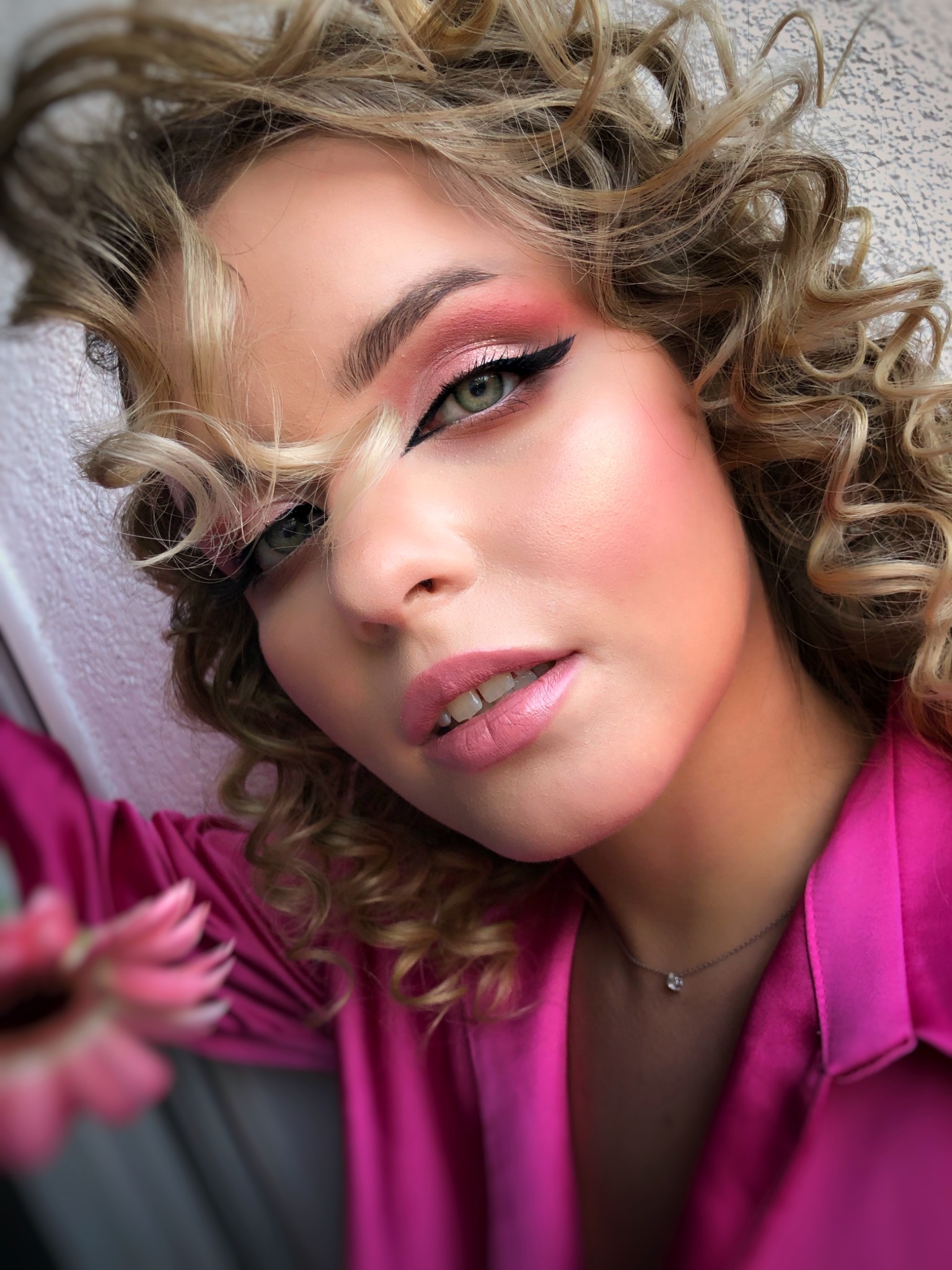 Portret kobiety z blond lokami, różowym makijażem oczu i ust, czarną kreską na powiece i delikatnym naszyjnikiem.