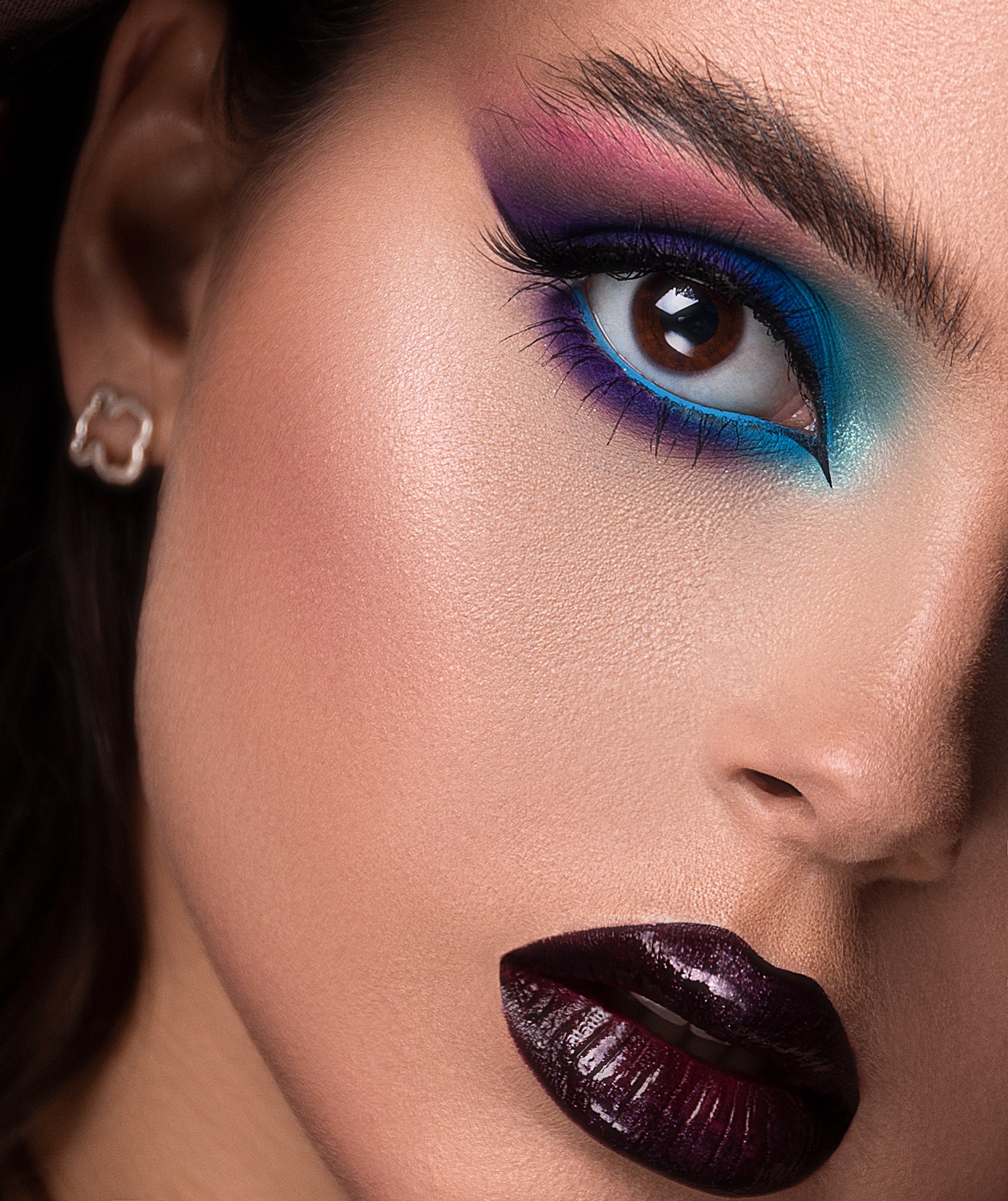 Portret kobiety z intensywnym, kolorowym makijażem oczu w odcieniach różu, fioletu i turkusu oraz ciemną, błyszczącą szminką w kolorze wina.