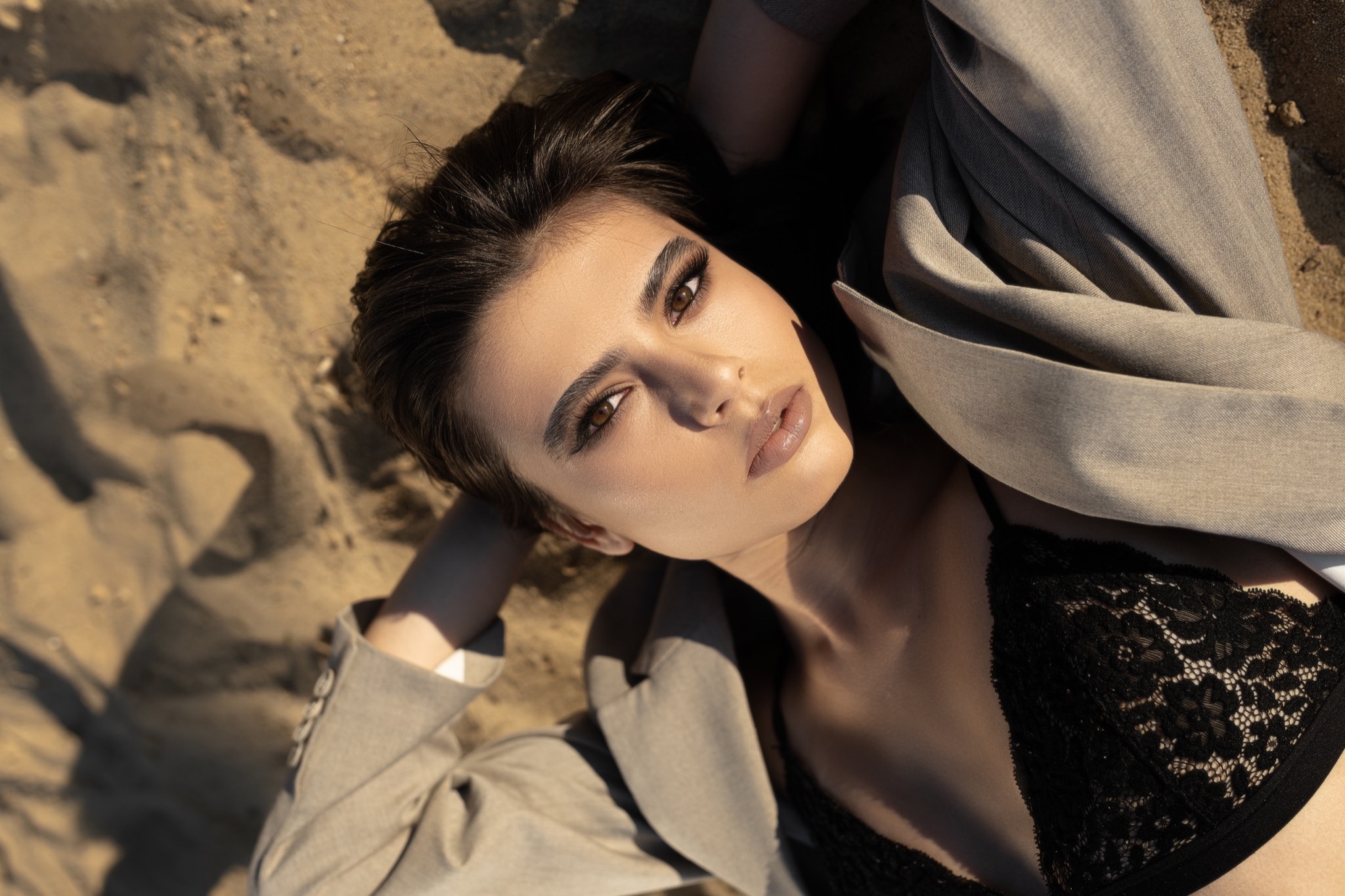 Portret kobiety leżącej na piasku, z wyraźnie podkreślonym makijażem oczu i ust, w stylizowanej sesji zdjęciowej z elementami mody.