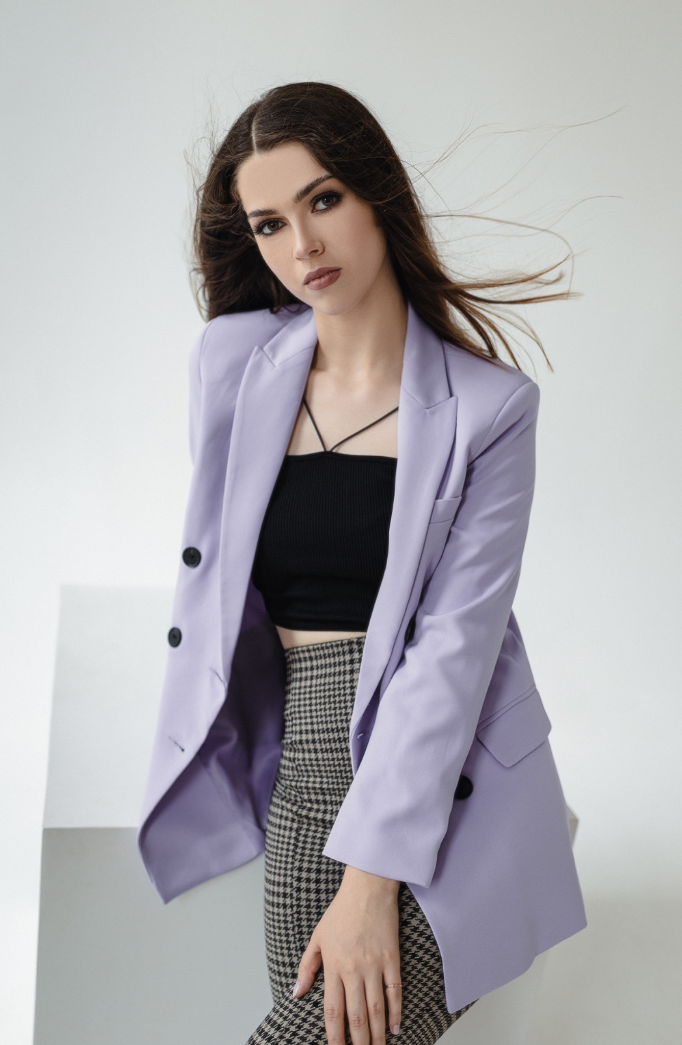 Portret kobiety z delikatnym makijażem oczu, podkreślonymi brwiami, w liliowym blezerze, czarnym topie i spódnicy w pepitkę, z rozwianymi włosami na jasnym tle.