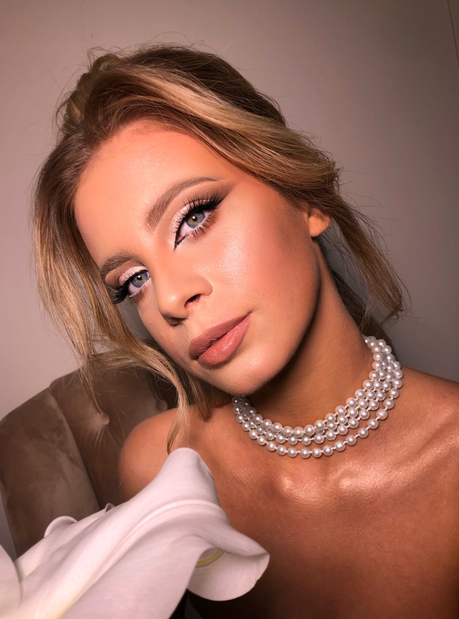 Portret kobiety z profesjonalnym makijażem oczu, podkreślonymi brwiami i błyszczącą cerą, ubrana w perłowy naszyjnik i trzymająca białą lilię.