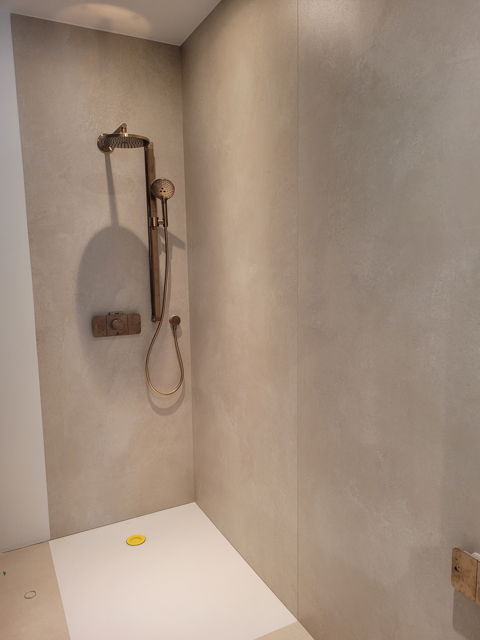 Nowoczesna kabina prysznicowa walk-in z deszczownicą w kolorze mosiądzu, minimalistyczny design, beżowe płytki imitujące beton, odpływ liniowy z żółtą zaślepką.