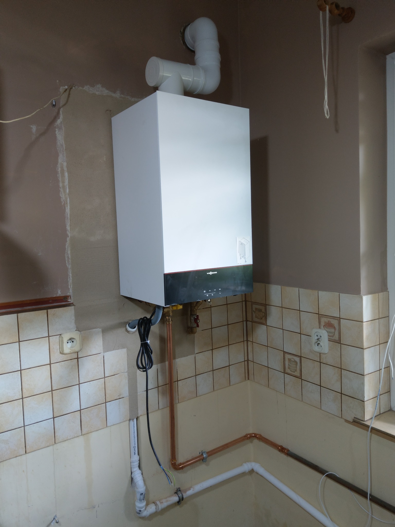 Nowo zainstalowany, biały piec gazowy Viessmann z czarnym panelem sterowania, podłączony do rur miedzianych i białych rur PCV w pomieszczeniu z kafelkami w odcieniach beżu i brązu, z widocznymi...