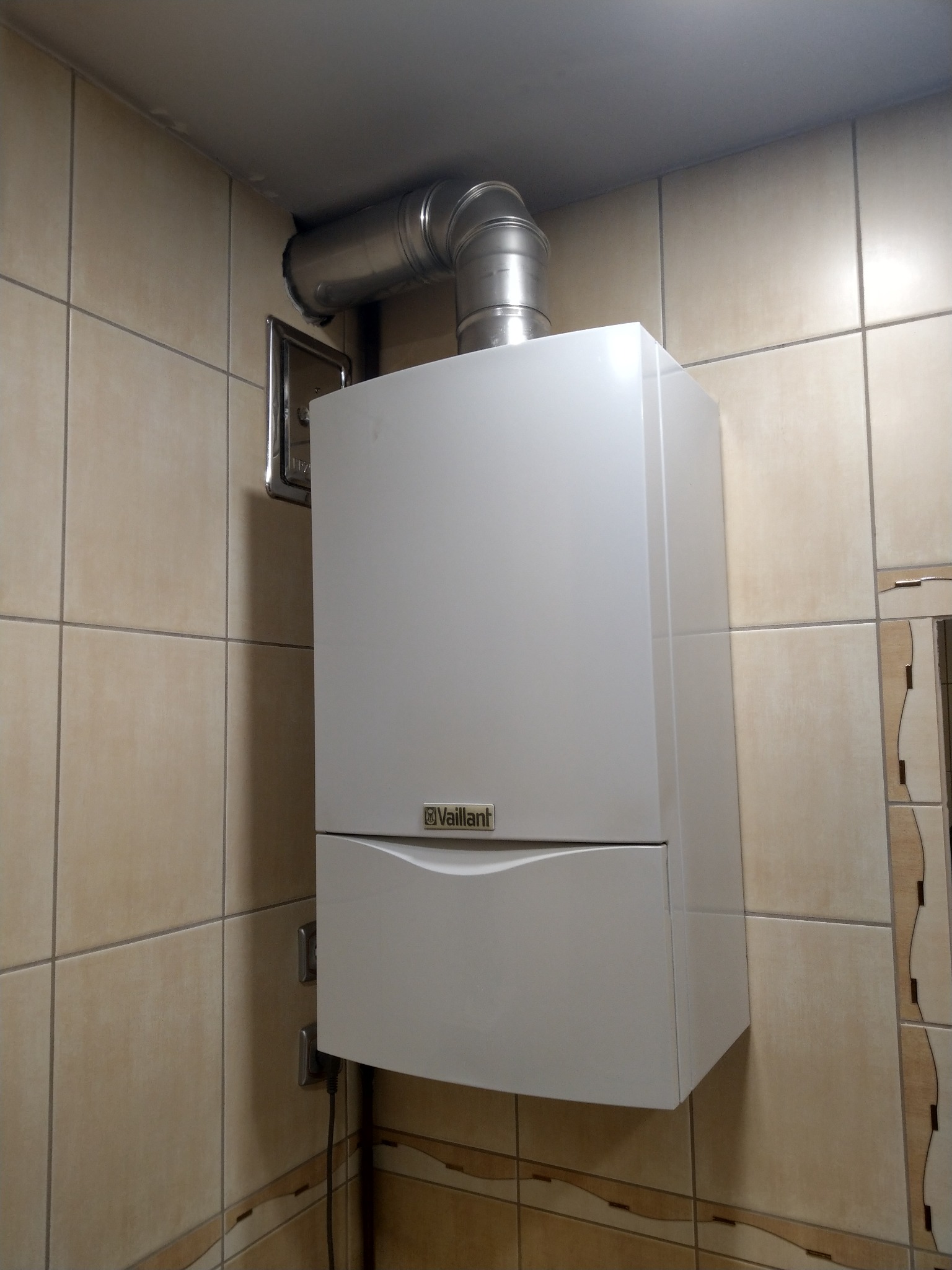 Biały wiszący piec gazowy Vaillant z metalowym kominem, zamontowany na ścianie wyłożonej beżowymi płytkami w łazience. Widoczne gniazdka elektryczne i dekoracyjne płytki na ścianie.
