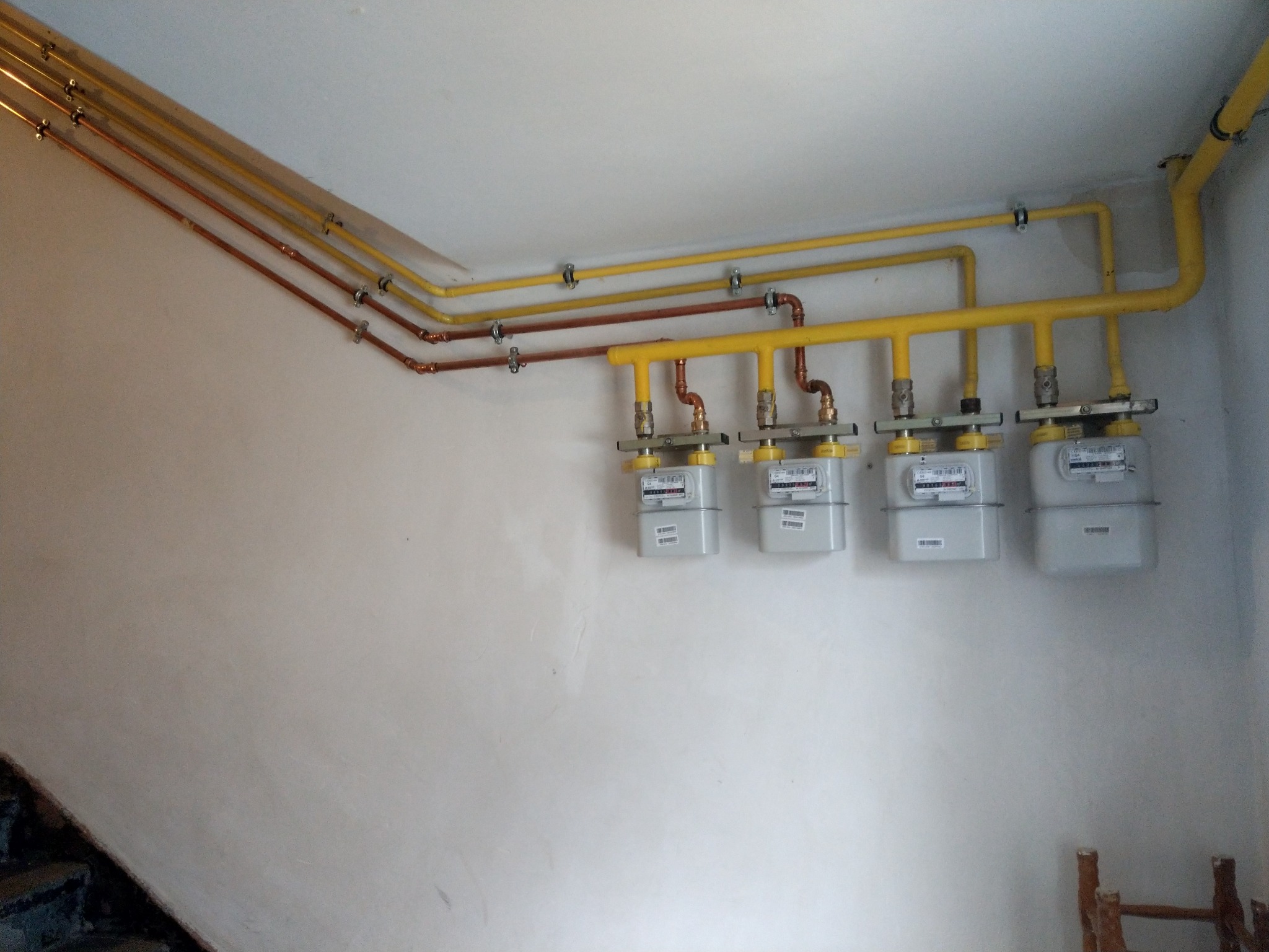 Cztery liczniki gazu zamontowane na ścianie, z rurami miedzianymi i żółtymi, widoczne elementy instalacji gazowej.