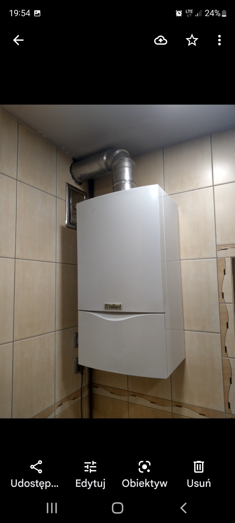 Biały, wiszący piec gazowy marki Vaillant z metalową rurą odprowadzającą spaliny, zamontowany na ścianie wyłożonej beżowymi płytkami w łazience.