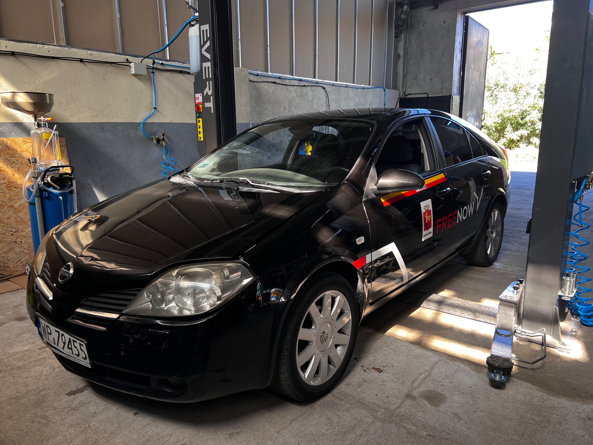 Czarny samochód Nissan Primera z naklejkami Free Now, zaparkowany w warsztacie samochodowym obok podnośnika i urządzenia do wymiany oleju.