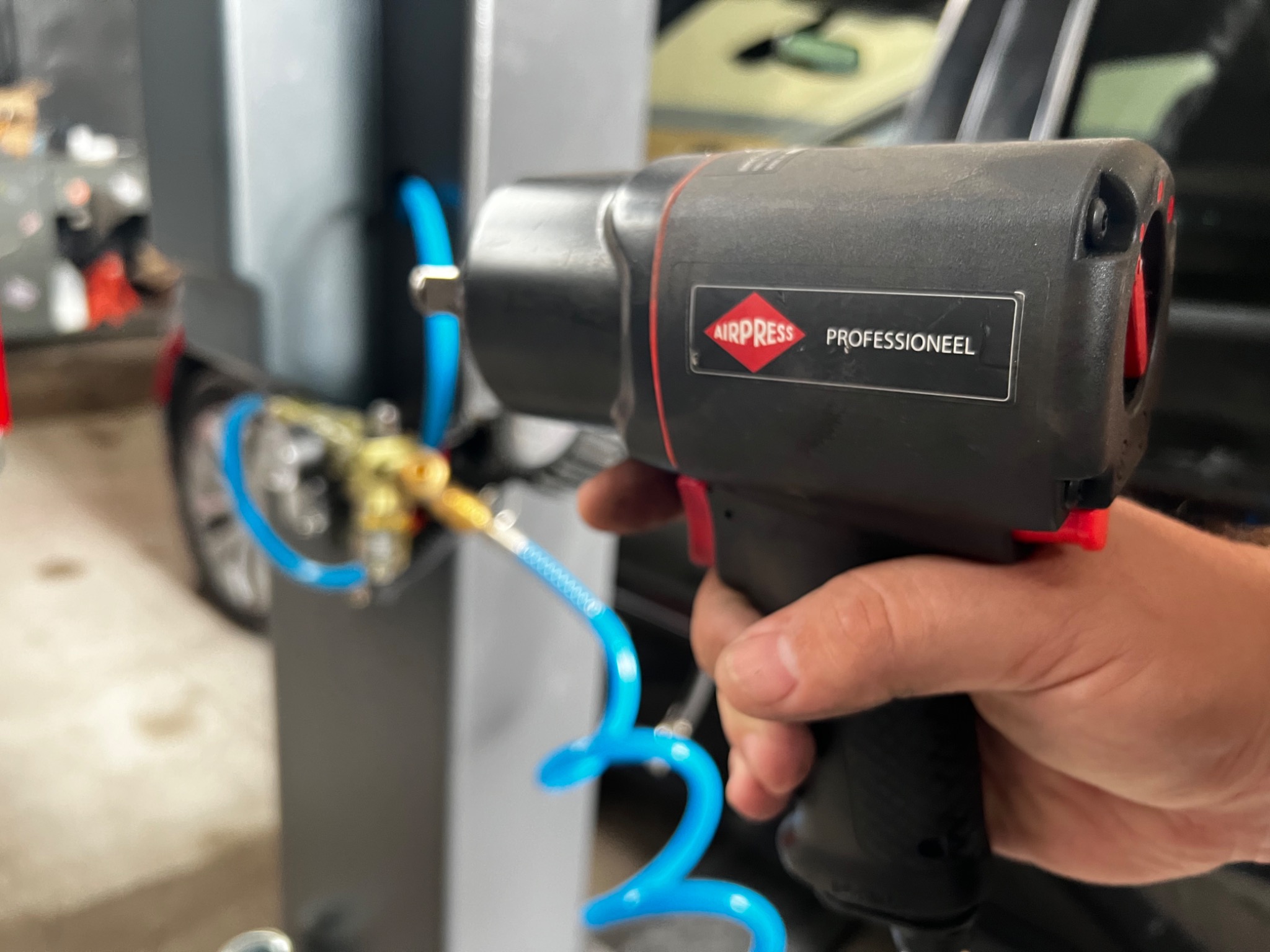 Pneumatyczny klucz udarowy marki Airpress Professioneel, podłączony do kompresora za pomocą niebieskiego węża spiralnego, trzymany w dłoni w warsztacie samochodowym.