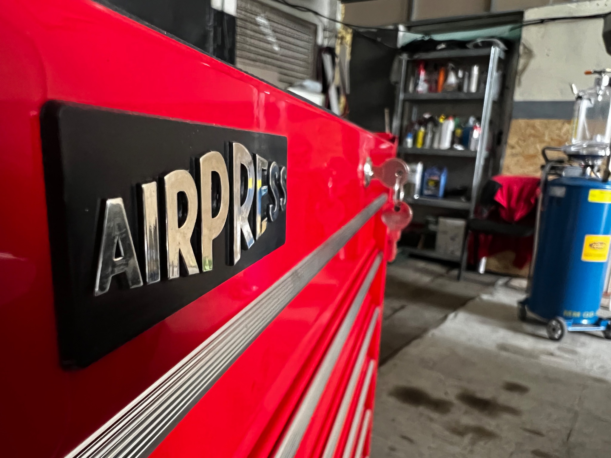 Czerwona szafka narzędziowa Airpress z chromowanymi detalami i kluczykami w zamku, w tle warsztat z regałem i niebieskim urządzeniem na kółkach.