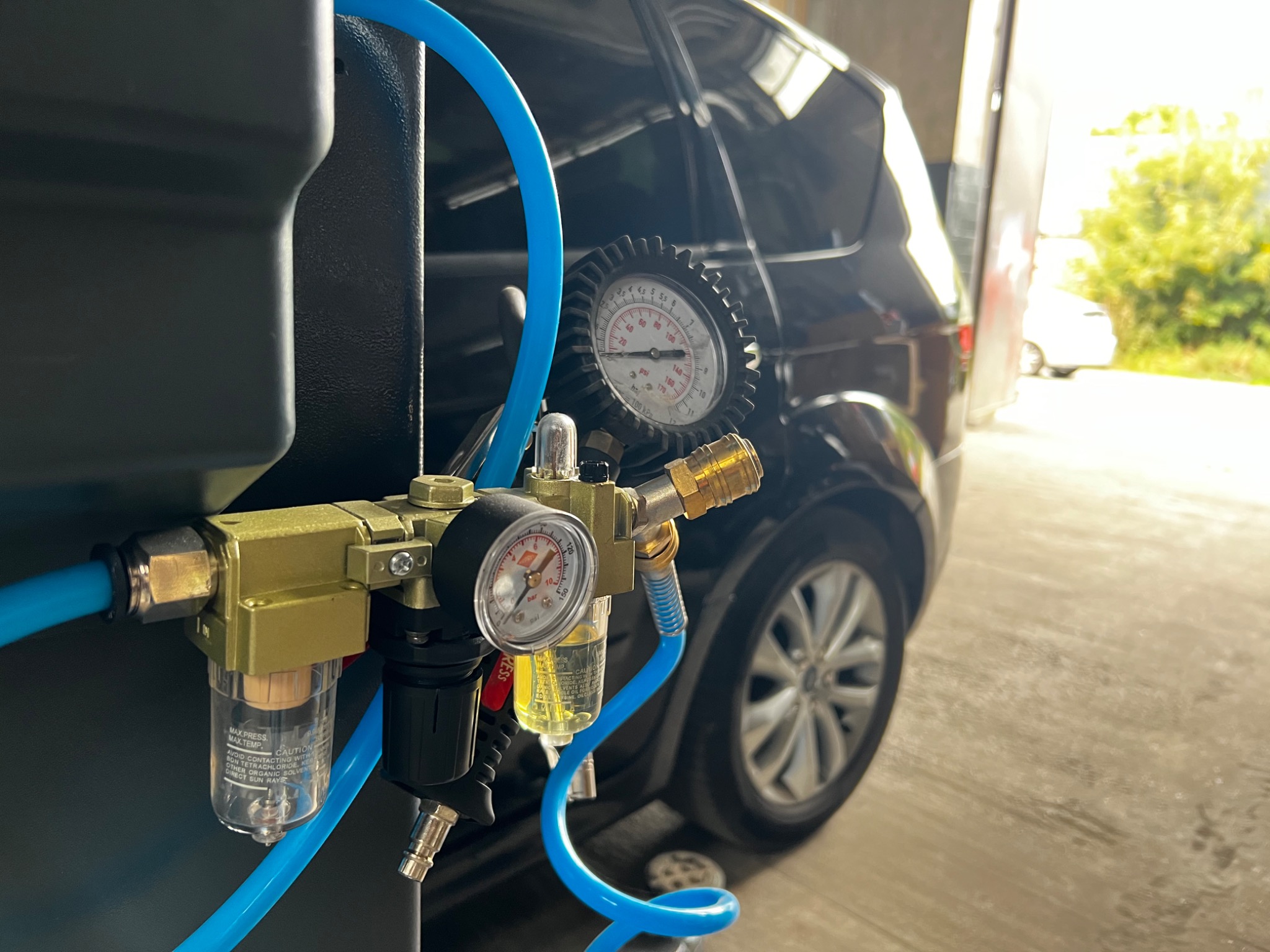 Sprzęt diagnostyczny z manometrami i filtrem, podłączony niebieskim przewodem, na tle czarnego samochodu w warsztacie.