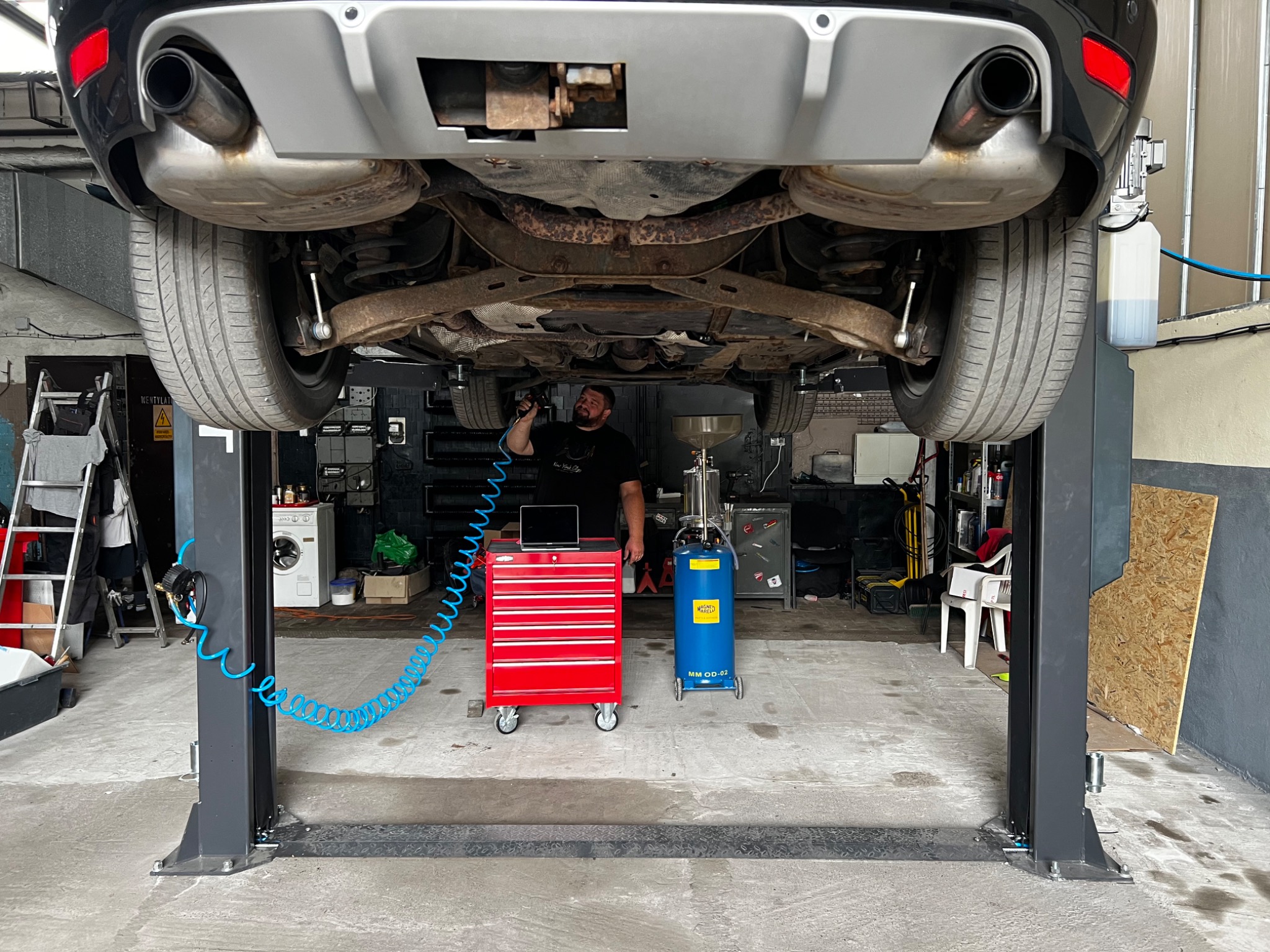 Czarny samochód na podnośniku w warsztacie, widok od spodu, mechanik z pistoletem pneumatycznym sprawdza podwozie, obok czerwony wózek narzędziowy i niebieski zbiornik na olej.