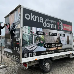 Pracownik na drabinie montuje okleinę reklamową na samochodzie dostawczym z hasłem 'Okna do Twojego domu' i zdjęciami okien, schodów i drzwi.