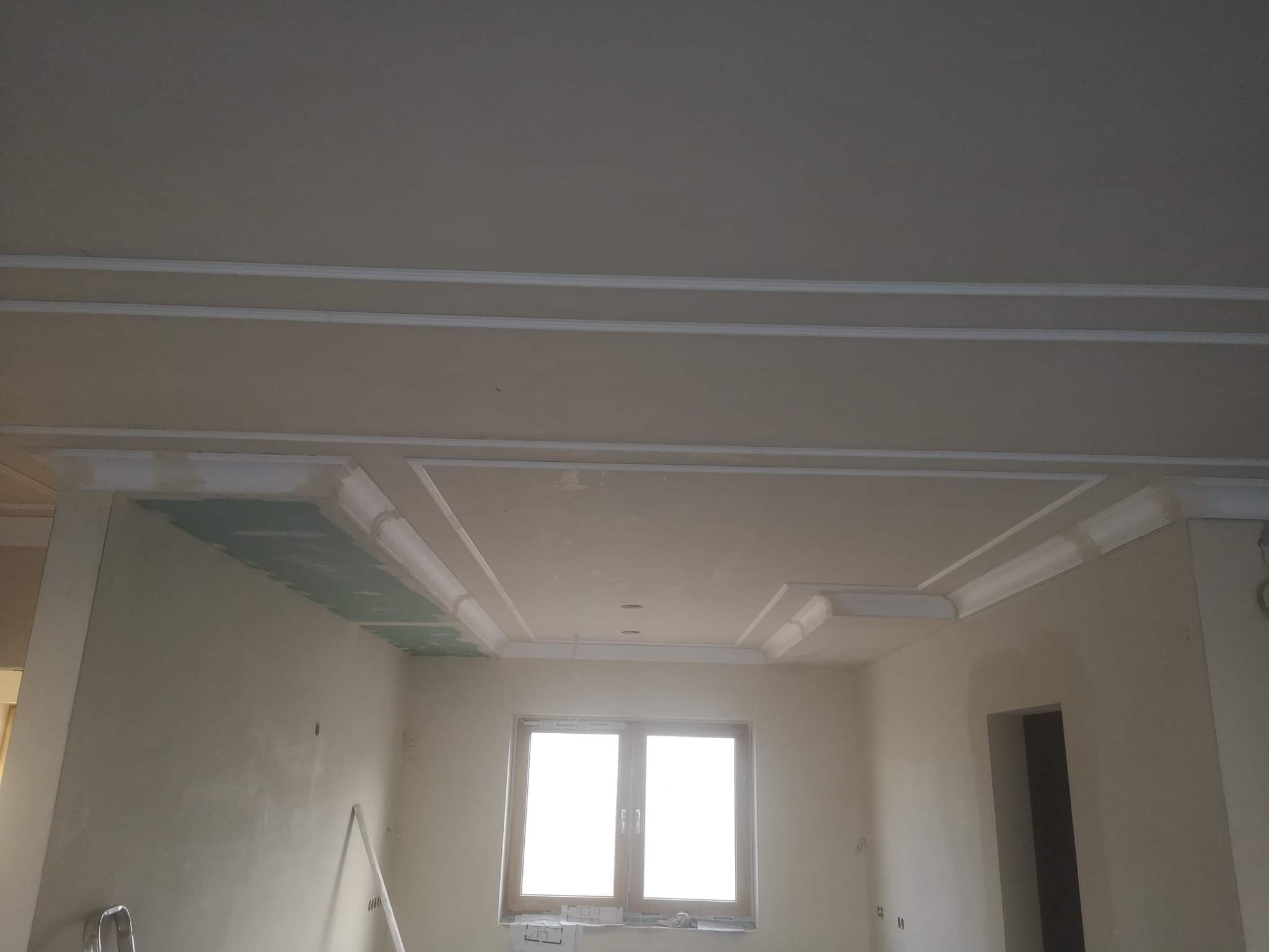 Pokój w trakcie prac wykończeniowych: surowe ściany, okno, ozdobne listwy sufitowe i fragment płyty gipsowo-kartonowej na suficie.