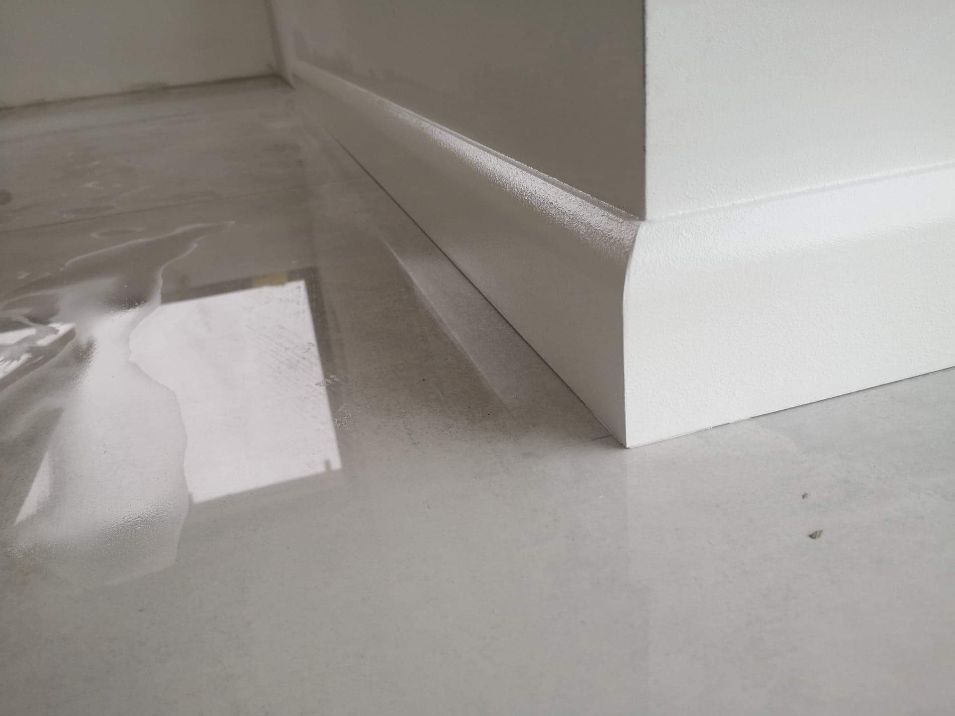 Świeżo pomalowana biała listwa przypodłogowa z widocznymi śladami wilgoci na błyszczącej, jasnej podłodze.