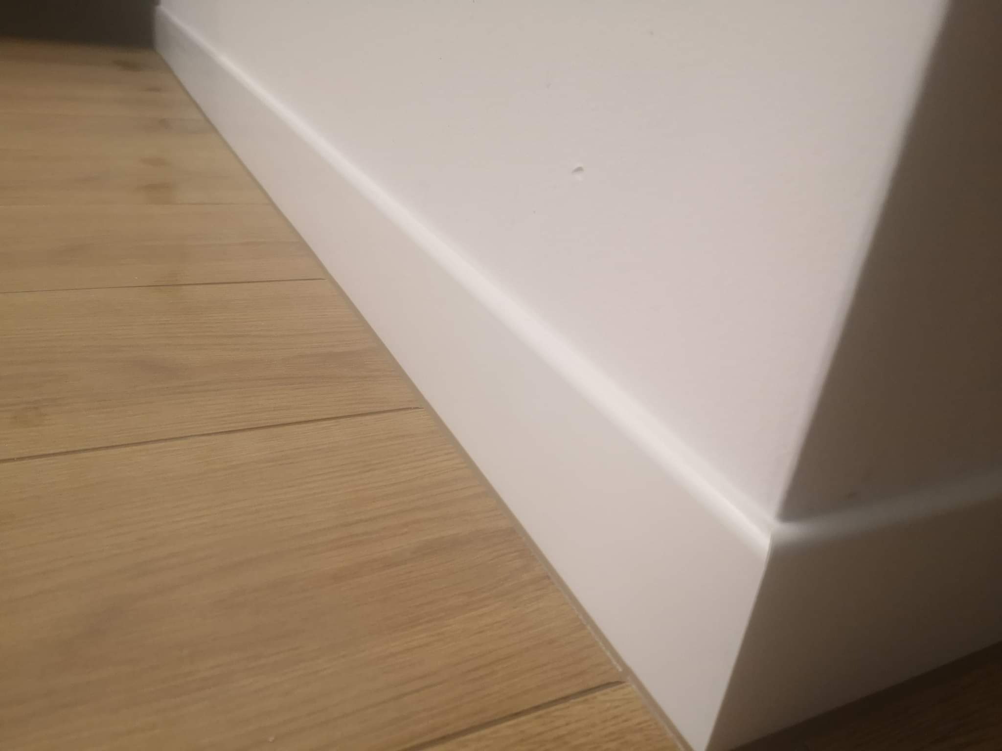 Fragment wnętrza: świeżo ułożona, jasna podłoga z paneli drewnianych i biała listwa przypodłogowa przy ścianie, widoczne połączenie paneli.