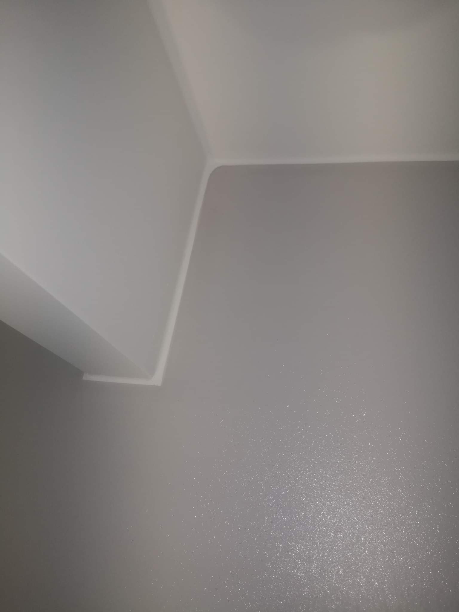Świeżo pomalowany narożnik sufitu i ściany w odcieniach szarości i bieli, widoczne delikatne refleksy światła na powierzchni ściany.