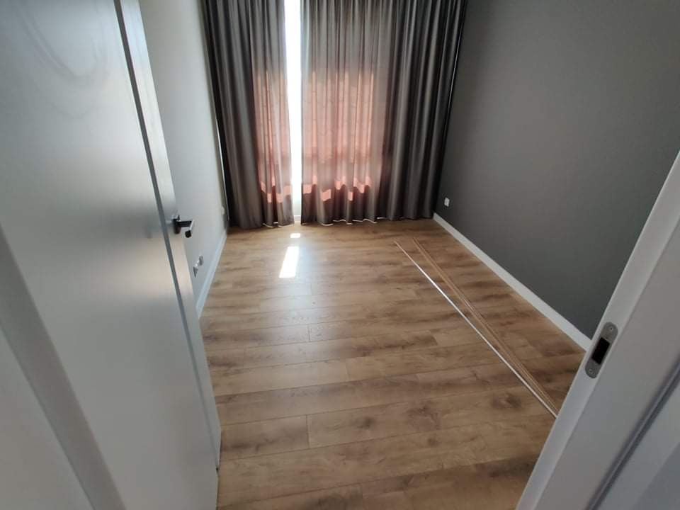 Wnętrze pokoju z ułożonym parkietem w odcieniach brązu, szare ściany, białe listwy przypodłogowe, zasłonięte okno z wpadającym światłem i białe drzwi po bokach kadru.